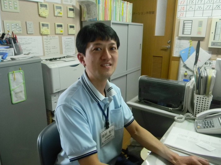 介護施設のスタッフが事務室で業務中。名札を着用し、デスク周りには書類や機器が配置されている。