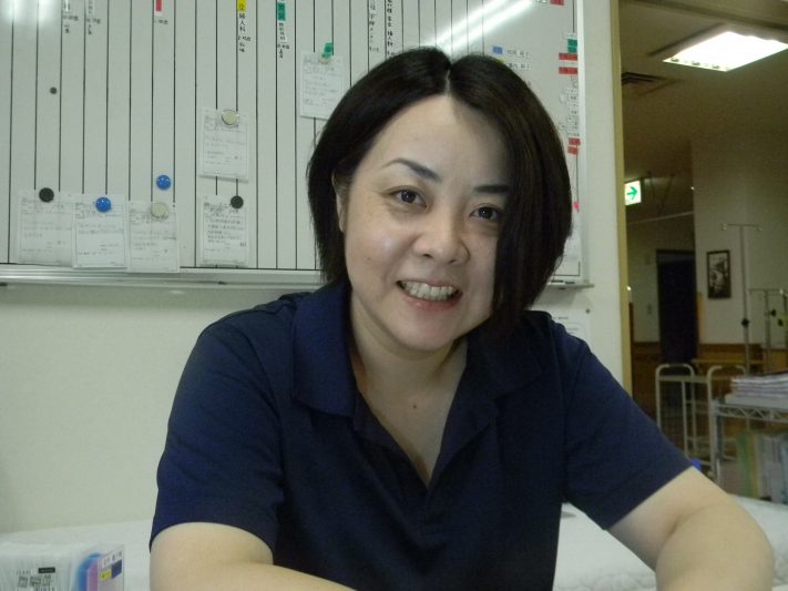 介護施設のスタッフが勤務室で笑顔を見せるポートレート。背後に業務用の掲示板が見える。